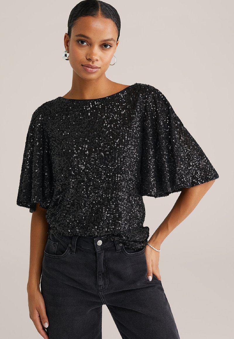 Top in paillettes nere con maniche a pipistrello, scollo rotondo e superficie texturizzata. Abbinato a jeans neri, con vita alta e design aderente.