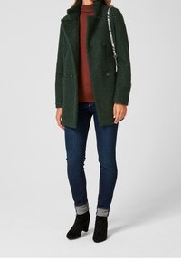 Mujer vestida con un abrigo texturizado verde oscuro, un cuello de tortuga óxido, jeans azul oscuro, botas negras hasta el tobillo y llevando un bolso tipo bandolera con estampado de piel de serpiente.
