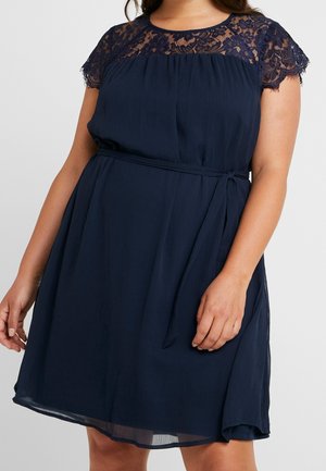 Vrouw draagt een knielange marineblauwe jurk met gebreide kapmouwen en een gestrikte tailleband.