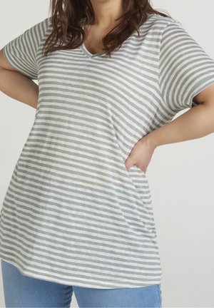 Vrouw in een T-shirt met korte mouwen, lichtgrijs en wit gestreept V-hals, met handen in de zij tegen een effen achtergrond.