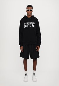 Μαύρη κουκούλα με το "VERSACE JEANS COUTURE" σε λευκή γραφή, συνδυασμένη με μαύρες βερμούδες και λευκά αθλητικά. Μοντέλο που φοράει γυαλιά ηλίου.
