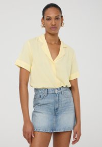Camicia gialla chiara a maniche corte con colletto, abbinata a una mini gonna in denim azzurro chiaro a vita alta. Texture liscia, stile casual.