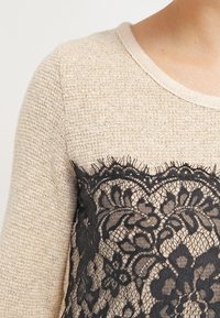 Pullover beige a maglia con una superficie testurizzata. Sovrapposizione in pizzo nero con motivi floreali, che mette in evidenza dettagli intricati nella zona del petto.