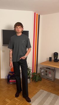 T-shirt cinza de manga curta com logótipo, calças pretas ajustadas e sapatos castanhos, contra uma parede riscada colorida numa sala.
