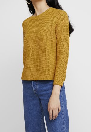 Mosterdgele gebreide trui met een ronde halslijn, geribbelde structuur en driekwart mouwen, gedragen met blauwe rechte jeans.