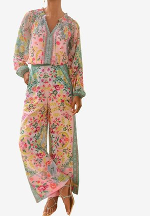 Femme portant un chemisier floral rose pastel à manches longues et un pantalon large avec un liseré vert, associé à des sandales beiges à brides.