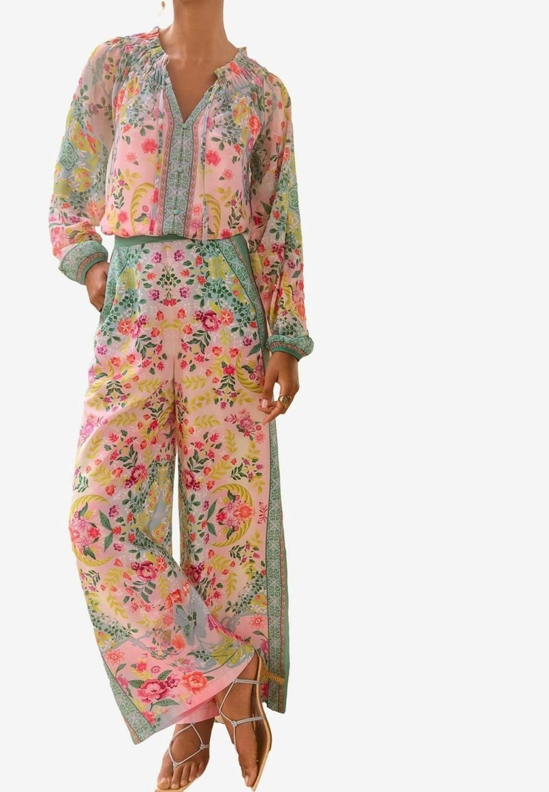Femme portant un chemisier floral rose pastel à manches longues et un pantalon large avec un liseré vert, associé à des sandales beiges à brides.