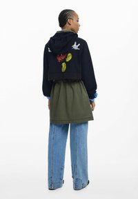 Blickdichter Navy-Hoodie mit grafischem Druck auf der Rückseite, kombiniert mit einem olivgrünen Rock, einem lockeren hellblauen Hemd und weit geschnittenen Jeans.