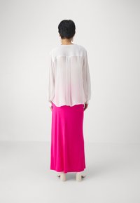 Hvit gjennomsiktig bluse med lange ermer og løs passform, parret med et lyst rosa, gulvlengde skjørt; modellen bruker lyse blokkheler.