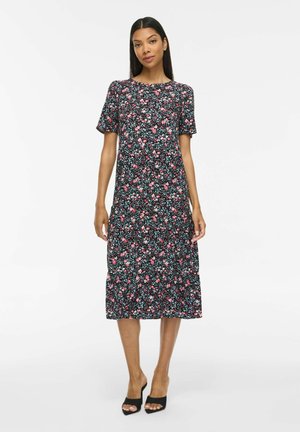 Femme portant une robe midi noire avec un imprimé floral rose et blanc et des manches courtes, associée à des talons ouverts noirs, debout sur fond blanc.