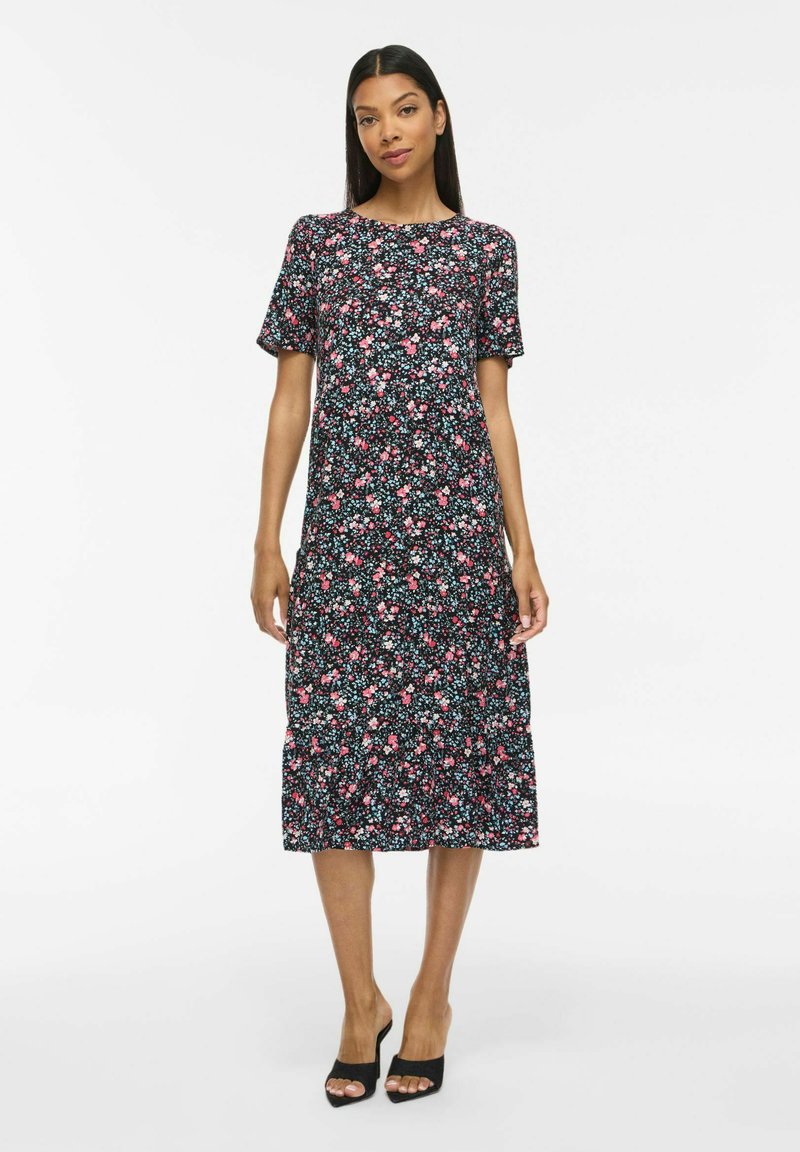 Femme portant une robe midi noire avec un imprimé floral rose et blanc et des manches courtes, associée à des talons ouverts noirs, debout sur fond blanc.