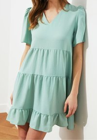 Mátově zelené šaty s krátkým rukávem, výstřihem do V, s tiered designem a volánkovým lemem. Vyrobeno z lehkého materiálu s jemnou strukturou.