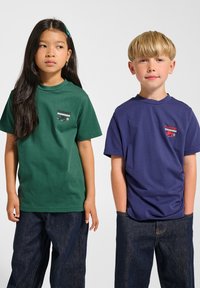 To børn iført t-shirts med korte ærmer: en grøn med et lille logo, en marineblå med et kontrastlogo, begge parret med blå denimjeans.
