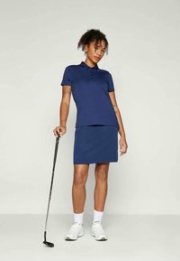 Námořnické polo tričko s límečkem, knoflíky a ladící úzkou sukni. Modelka drží golfovou hůl, obléká bílé tenisky a ponožky.