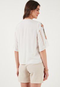 Top blanco ligero con mangas cortas, que presenta bordados coloridos en los hombros. La parte trasera tiene un ajuste relajado y una textura forrada.