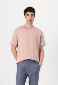 Hellrosa T-Shirt mit kurzen Ärmeln aus weichem Stoff, kombiniert mit grauen, gefalteten Hosen, die einen klassischen Rundhalsausschnitt und eine lockere Passform aufweisen.