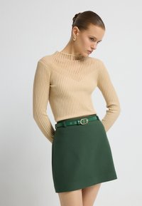 Beige ribbeltrui met lange mouwen en hoge hals, gecombineerd met een groene minirok en een bijpassende riem met een ronde gesp.