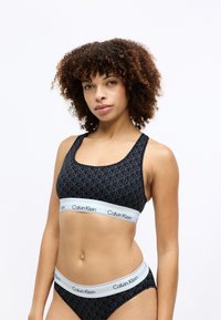 Sort sportsbh med geometrisk mønster, bred elastisk bånd med "Calvin Klein" branding i lyseblå, matchende sort bikiniunderdel.