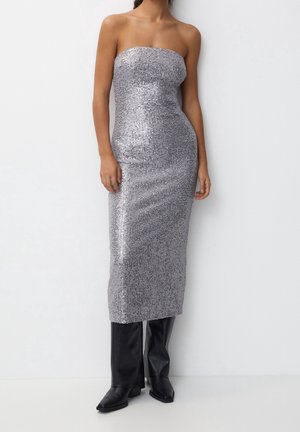 Femme portant une robe midi argentée à sequins sans bretelles, associée à des bottes en cuir noires montant jusqu’aux genoux, debout devant un fond blanc uni.