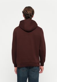 Brown hooded sweatshirt with a textured finish. Features a standard cut, ribbed cuffs, and hem. Worn with blue jeans, shown from the back.