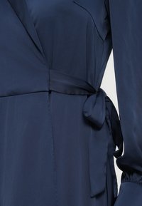 Marineblå omslagskjole laget av glatt stoff med knytebelte i midjen og lange ermer, med en subtil glans og lett leggdetalj.