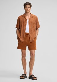Korte mouwen rustkleurig shirt met een gestructureerd patroon, gecombineerd met bijpassende rustkleurige shorts. Zwarte platte sandalen maken de outfit af.