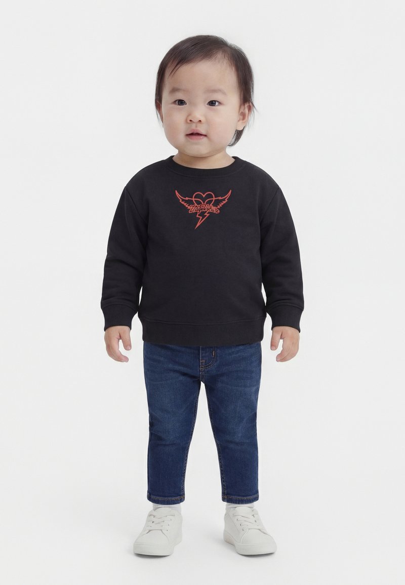 Μικρό παιδί όρθιο, φοράει μαύρο φούτερ με κόκκινη καρδιά και λογότυπο αστραπής, μπλε τζιν και λευκά αθλητικά παπούτσια σε λευκό φόντο.