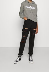 Felpa grigia con cappuccio e testo bianco, abbinata a pantaloni cargo neri rovinate con diverse strappi. Sneakers alte nere completano l'outfit.