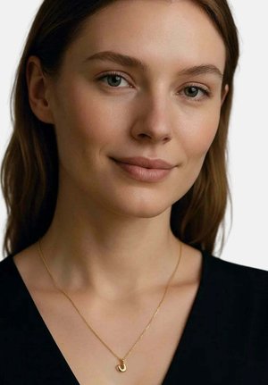 Jeune femme à la peau claire et aux cheveux bruns souriant légèrement, portant un haut noir et un collier pendentif en forme de fer à cheval en or.