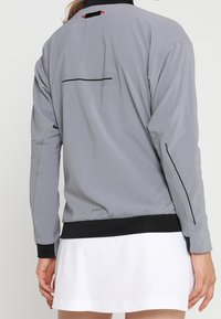 Grå lättviktsjacka med svarta detaljer. Har rund halsringning, dragkedjeficka på ryggen samt elastiska ärmslut och nederkant. Slät yta.