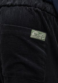 Calças de corduroy pretas com riscas verticais, apresentando um rótulo verde com a inscrição "SCOTCH & SODA" no bolso esquerdo.