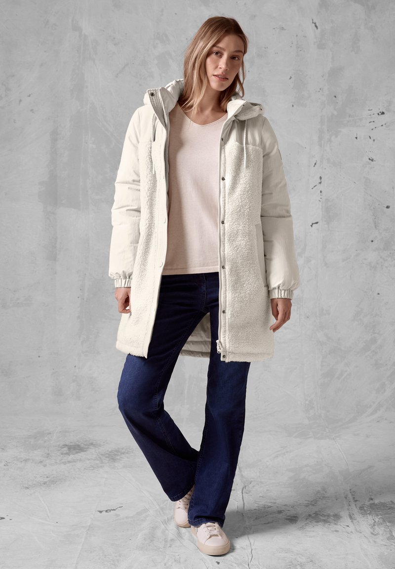 Lange witte pufferjas met fleecepanelen, elastische manchetten en een capuchon. Gdragen over een lichtroze shirt en donkerblauwe jeans met witte sneakers.