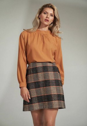 Bluzka z długim rękawem w kolorze rdzy z plisowanymi detalami, połączona z plisowaną spódnicą w kratę w czarno-brązowo-pomarańczowe wzory.