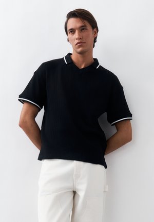 Νεαρός άνδρας με σκούρα μαλλιά, φοράει μαύρο κοντομάνικο πουκάμισο με λευκή τρέσα και λευκό παντελόνι, στέκεται μπροστά σε λευκό φόντο.