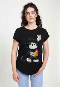 Černé bavlněné tričko s potiskem Mickey Mouse oblečeného do šortek s multibarevným duhovým vzorem a gestem znamení míru.