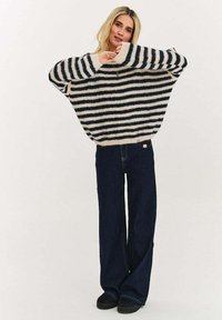 Přehnaně velký pruhovaný svetr v námořnické modři a krémové barvě, s širokými rukávy a měkkým, texturovaným úpletem. Kombinován s tmavými džínovými rozšířenými kalhotami.