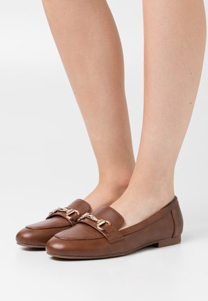 Mocassins en cuir marron avec une semelle plate, un bout arrondi et un accent de chaîne en métal sur le devant. Texture lisse avec des coutures minimales.