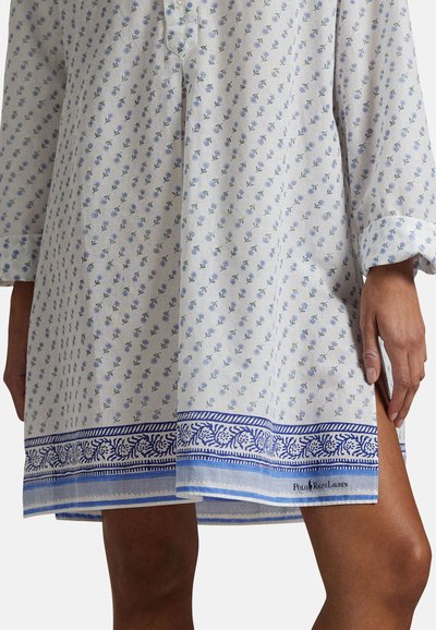 Femme portant une robe blanche avec de petites fleurs bleues imprimées et une bordure décorative bleue près de l'ourlet, montrant des jambes nues et une main.