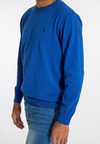 Blå stickad tröja med rund halsringning, ribbade ärmslut och nederkant. Har en liten, mörk logotyp på bröstet. Fabrik av bomullsblandning.