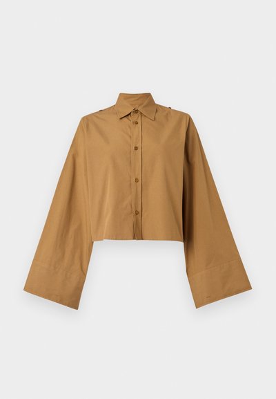 Klädd knapp-up skjorta i beige bomull, med spetsig krage och vida ärmar med en mjuk textur. Två knappar på axeln som accent.