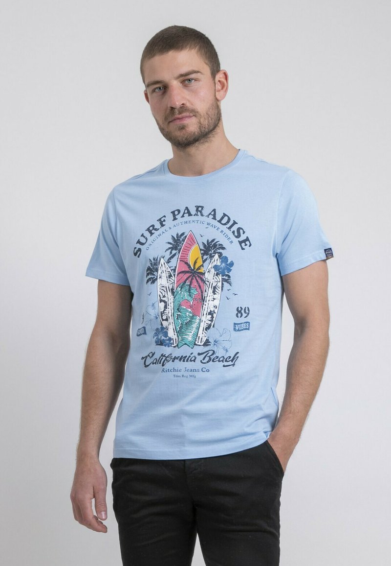 T-shirt en coton bleu clair avec une impression graphique comprenant des planches de surf, des palmiers et le texte "Surf Paradise". Manches courtes et col rond.