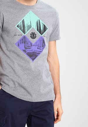 Personne portant un t-shirt gris avec un imprimé géométrique de forêt bleu et violet et un pantalon bleu foncé sur un fond clair uni.