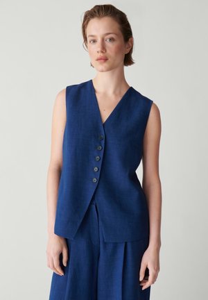 Frau trägt eine ärmellose marineblaue Weste mit Knöpfen und passende weite Hosen vor einem einfarbigen grauen Hintergrund.
