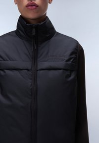 Schwarze gepolsterte Weste mit hohem Kragen, Reißverschluss und Taschen auf der Vorderseite. Das Material wirkt glatt mit einer feinen Textur. Logo auf der Brust.