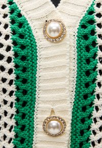 Πλεκτή ζακέτα με πράσινο και εκρού μοτίβο και υφή πλέξης πλέγματος. Διακρίνονται δύο μεγάλα κουμπιά από πέρλες διακοσμημένα με στρας.