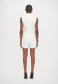 Vit ärmlös overall med skräddarsydd design, med stängd krage och korta shorts; ihop med svarta klackskor.