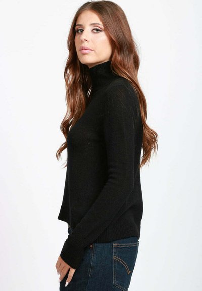 Donna con lunghi capelli castani mossi, che indossa un maglione a collo alto nero e jeans blu scuro, in piedi di lato su uno sfondo bianco.