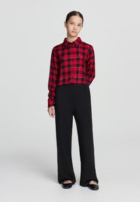 Camicia a quadri rossa e nera con bottoni e maniche lunghe, abbinata a pantaloni larghi neri e scarpe flat nere, su uno sfondo semplice.