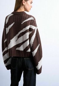 Femme portant un pull tricoté court marron avec des rayures abstraites blanches et un jean taille haute noir, vue de dos et de côté.