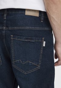 Jeans de mezclilla azul oscuro con un parche de cuero con texto en la cintura. Tienen costuras amarillas en contraste y dos bolsillos traseros.